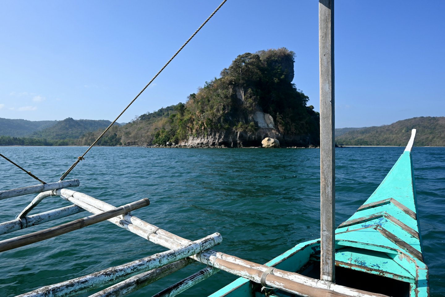 Turquoise boat with bamboo outriggers approaches a tropical island.