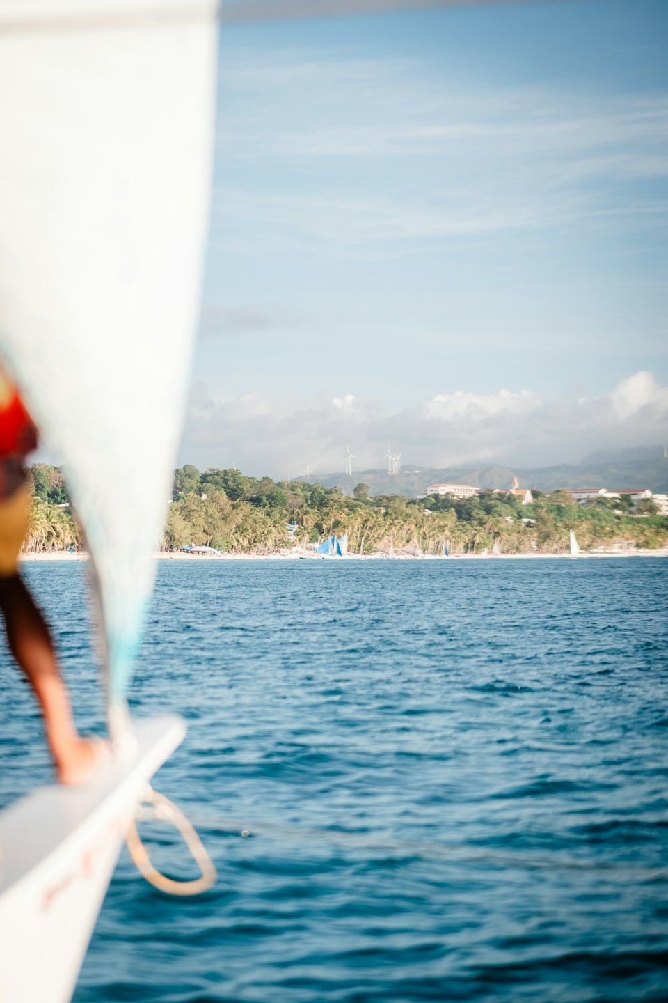 Sailboat traveling toward a tropical island with palm trees and wind turbines.