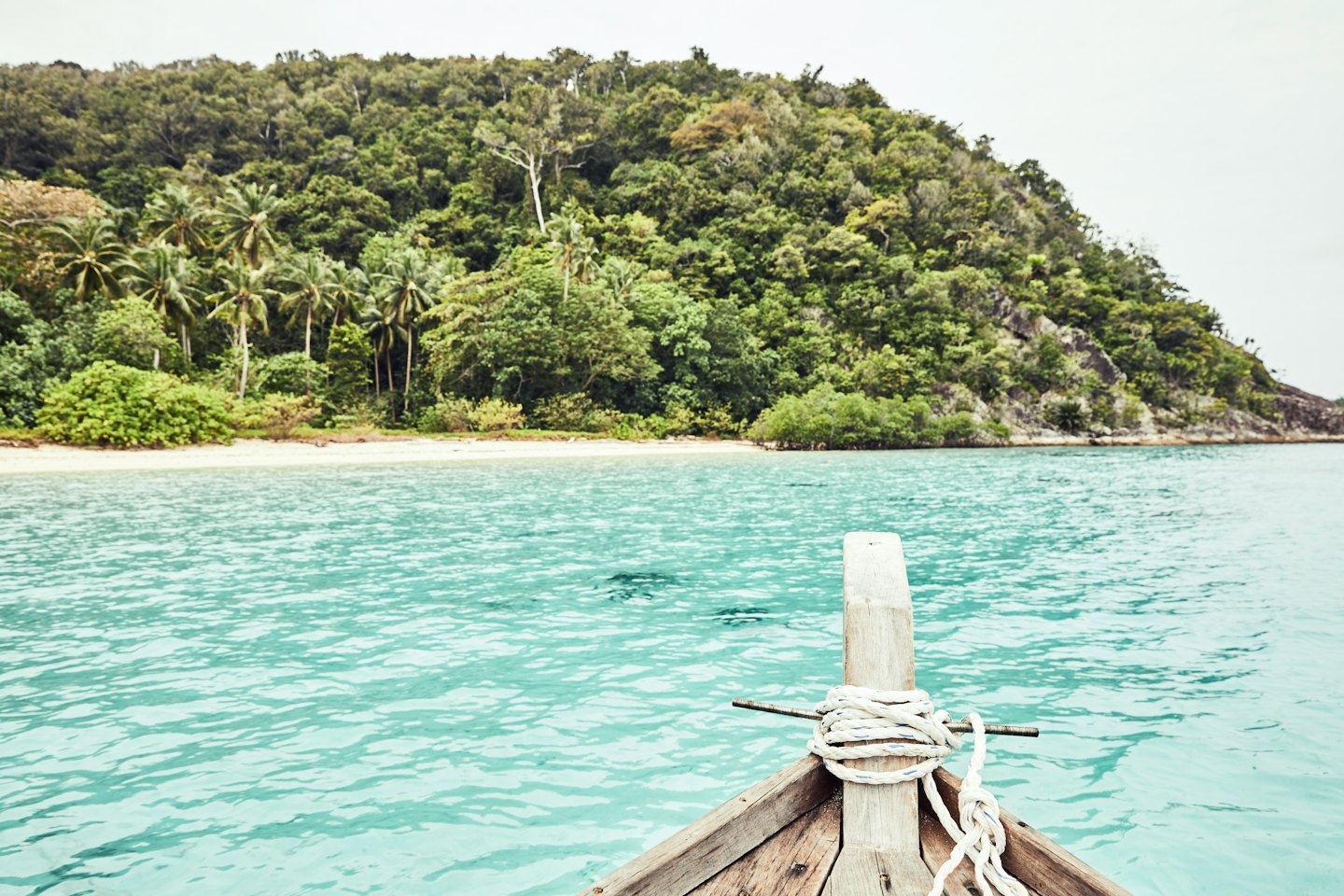 Wooden boat bow sailing toward a sandy tropical island beach.