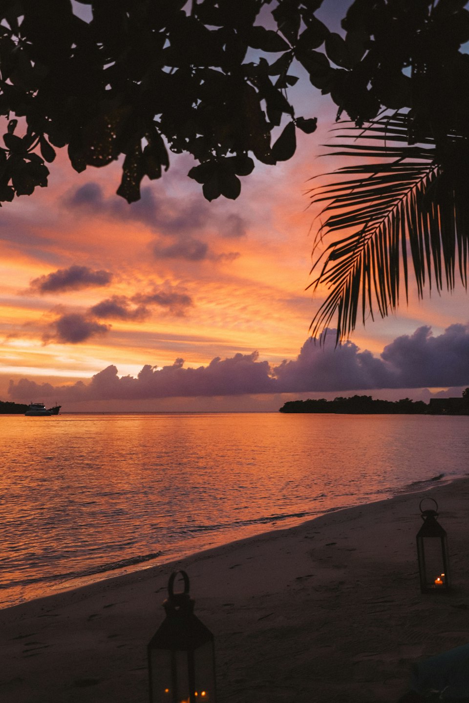 Two lit lanterns sit on a sandy beach at sunset.